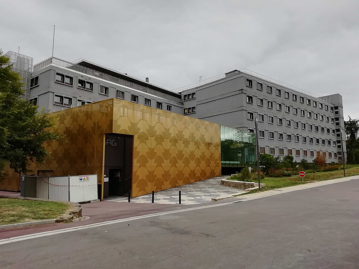 Das Foto zeigt die Maison du Portugal. Das Haus besteht aus zwei parallel gesetzten grauen Baukörpern. Im Vordergrund befindet sich ein moderner gold-brauner Anbau, der als Eingangsbereich dient. 
