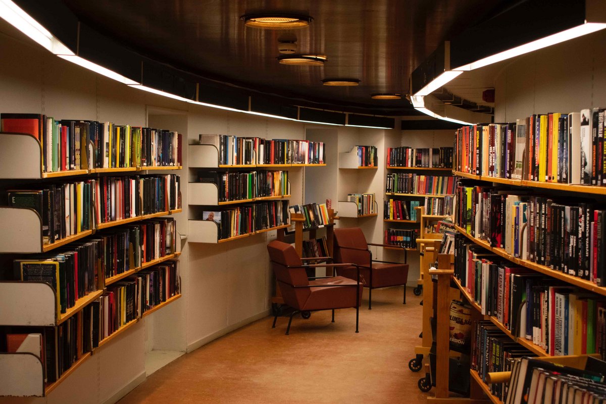 Das Foto zeigt einen schwach beleuchteten Gang in einer Bibliothek. An den Wänden sind Bücherregale. Zwei Sessel laden zum Schmökern ein.